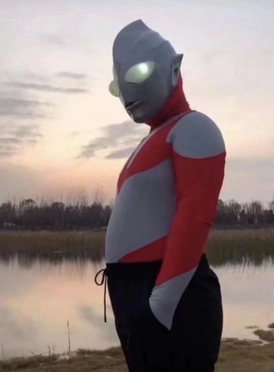 图片中显示一个穿着奥特曼服装的人站在湖边，背景是美丽的夕阳。这个人穿着经典的红银相间的奥特曼上衣，但下身却搭配了一条普通的黑色运动裤，形成了一种有趣的反差感。奥特曼标志性的发光眼睛清晰可见，他双手插在裤袋里，侧身望向远方，表情平静。湖边的水面倒映着天空的晚霞，远处有一排树木，整个场景营造出一种宁静而又略带幽默的氛围。这种将经典特摄角色与日常休闲元素结合的方式，创造出了一种滑稽又接地气的视觉效果。