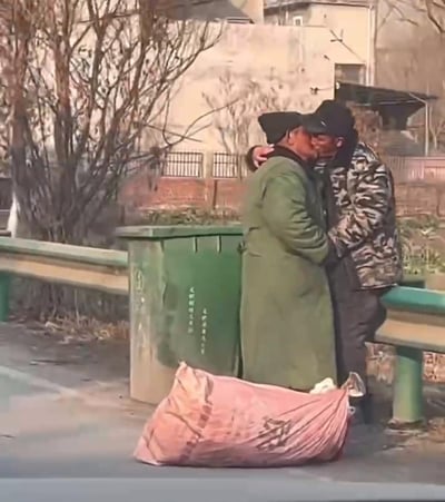 在城郊或乡村的路边，一位身着军绿色长款棉服的老人与一位穿迷彩棉服的老人紧紧相拥，姿态亲昵，旁边地面放着装物品的蛇皮袋，背景是绿色垃圾桶、护栏和枯树，整体环境朴实。画面传递出一种平凡又真挚的温暖，没有刻意的修饰，满是生活里的人情味。
