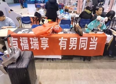 这是一张线下展会现场的实拍图，场景为室内的同人/兽展活动场地，画面核心是摊位前悬挂的橙色横幅，上面写着白色大字“有福瑞享 有男同当”，是对俗语“有福同享，有难同当”的谐音改编：“福瑞”指代Furry（兽人爱好者群体），“男同”指代男同性恋群体，用趣味化的谐音梗将两个群体关联起来，吸引同好关注。摊位上摆放着橙色收纳袋、兽装头套、随身包等物品，周围有多位参与活动的人群，前景处还有一个黑色拉杆行李箱，整体氛围轻松热闹，是亚文化爱好者线下聚集的场景。