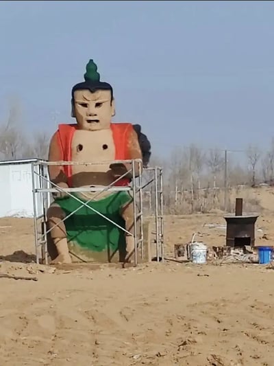 图片展示一尊用土堆做成、类似佛像的大型雕塑，现场搭有施工用的脚手架，还能看到旁边有取暖炉、油漆桶等物品，画面背景荒凉且有些简陋。由于造型憨态，场景荒诞，使人联想到网友调侃的“临时工程”、“佛系”梗等，具有一定的搞笑效果和现实暗示感。佛像的造型略显粗糙，色彩搭配有点跳脱，给人一种“草率但认真”、“手工艺人版佛像”的感觉。该场景容易引发网友关于乡村、施工队或短视频创作的吐槽。

如果要分析双关或暗示：整体画面隐含着对现实生活“佛系”、“临时抱佛脚”这种文化现象的调侃。