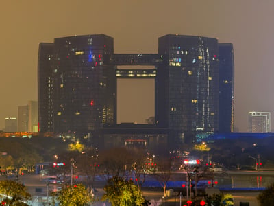 这是一张杭州地标建筑的夜景实拍图，画面中深色的摩天大楼带有巨大的方形镂空结构，楼体的灯光在夜晚中零星亮起，背景是朦胧的灰黄色夜空，前景是带有路灯和树木的广场区域。发布者认为该场景的冷峻建筑风格与夜晚的氛围感，和游戏《赛博朋克2077》里荒坂塔附近的广场高度相似，充满赛博朋克的冷峻质感。