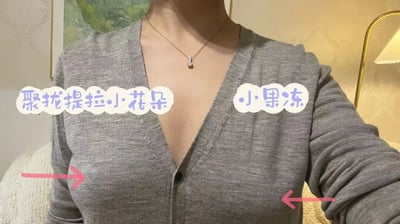 图片展示了一位穿着灰色开衫的女性上半身，重点展示了胸部区域。她佩戴着一条银色项链，背景中可以看到一盏金色台灯和部分绿色画作。图片上有两个文字标注：左侧标注为“聚拢提拉小花朵”，右侧标注为“小黑冻”，分别用粉色箭头指向胸部的不同位置，似乎在展示某种产品效果或服装特点。