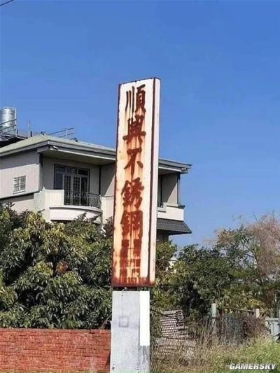 这是一张户外实拍图，画面主体是一块立式商家招牌，上面写有繁体的“順興不銹鋼”字样，意为顺兴不锈钢，是一家经营不锈钢相关业务的商家。但这块招牌本身已经严重生锈，大片红棕色锈迹覆盖了招牌下半部分，尤其是“不锈钢”三个字的位置锈迹格外明显，与商家经营的不锈钢不易生锈的属性形成强烈的反差讽刺效果。招牌后方是一栋两层居民楼，周围生长着茂密的绿植，底部有红砖墙，背景是晴朗的蓝色天空，整体氛围明亮，更加凸显了招牌生锈的违和搞笑感。