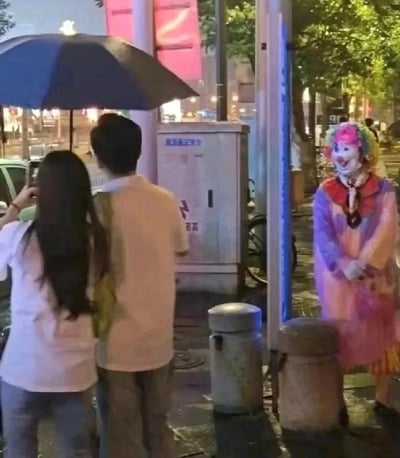 深夜的城市街道上，一对情侣撑着蓝色雨伞，背影面向一位穿着鲜艳彩色服装的小丑。小丑戴着五彩假发，脸上画着夸张的笑容，安静地站在人行道旁，背景有路灯、树木和模糊的城市灯光，营造出一种既诡异又略带荒诞的氛围。