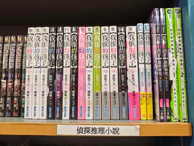 书店或图书馆的书架局部特写，主要展示了多本《我推的孩子》漫画单行本整齐排列，从第2卷到第16卷（其中11、12卷缺失），封面以黑白为基调，部分卷册带有粉、绿等颜色点缀，作者均标注为赤坂明。左侧可见《BAKUMAN》等其他漫画系列，右侧有《我在意的孩子不是男人》等作品。书架下方标签明确写着‘侦探推理小说’，但实际陈列的《我推的孩子》等漫画多为青春偶像、娱乐圈题材，形成分类标签与内容类型的有趣错位。
