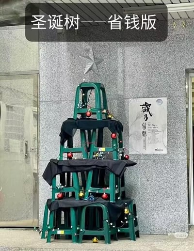 这是一个用绿色塑料凳子堆叠打造的「省钱版圣诞树」，一共四层由下到上凳子数量递减，每层凳子上都铺有黑色布料，用来模拟圣诞树的枝叶轮廓，同时搭配了红色圣诞球、金色小铃铛和白色串灯作为装饰。背景的灰色石质墙面上，刚好有一颗银色五角星，完美充当了圣诞树的顶星。图片上方有黑色文字标注「圣诞树——省钱版」，右侧还贴有一张写着「歲月留聲 12/14」的海报。整体是利用日常常见的塑料凳子DIY的趣味圣诞装饰，充满接地气的巧思，主打低成本打造节日氛围，创意十足又不失趣味，非常适合预算有限的节日布置参考。
