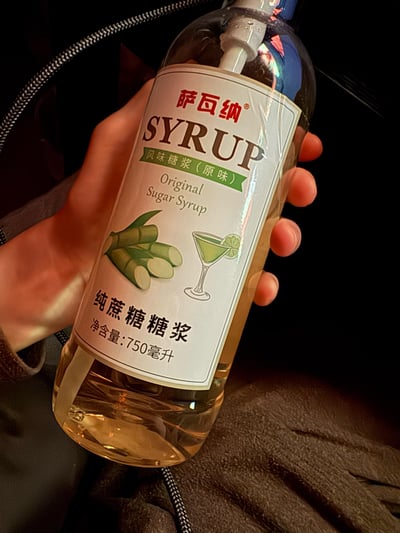 一只手握着一瓶750毫升的萨瓦纳原味纯蔗糖风味糖浆，瓶身标签清晰标注了品牌、产品类型、英文名称，还配有甘蔗原料图案和调酒杯示意图案，明确这是一款用于调制饮品的纯蔗糖糖浆，适合为咖啡、鸡尾酒等增添甜味。