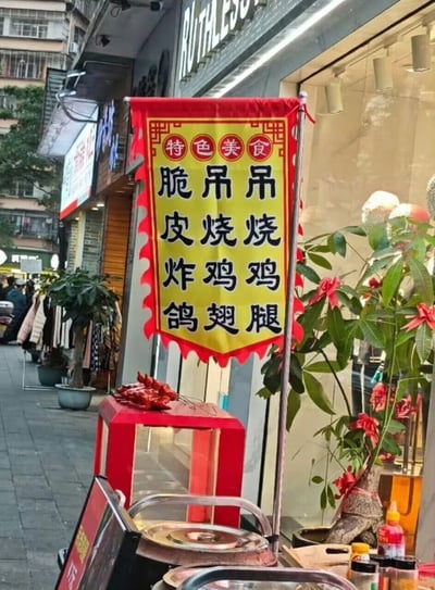 图片展示了一条热闹的街道场景，街边店铺门口悬挂着一面醒目的黄色红边横幅。横幅顶部写有“特色美食”字样，下方竖排列出“脆皮吊吊 皮烧烧 炸鸡鸡 鸽翅腿”等菜品名称，字体为黑色加粗，整体设计具有浓郁的中式传统广告风格。横幅下方摆放着红色的展示柜，里面可能陈列着食品样品，周围有绿植点缀，背景中可见其他商铺和行人，充满生活气息。