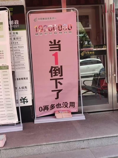 这是拍摄于中国邮政储蓄银行线下网点门口的宣传海报，海报以数字“1”隐喻健康，后续的“0”对应健康之外的金钱、房产、事业等生活要素，用醒目的标语“当1倒下了，0再多也没用”传递“健康是一切的基础”的理念，以此推广银行的健康类金融保险产品。画面左侧可见邮政网点的营业时间、业务服务信息、保险金额参考表等标识，背景是银行的玻璃门入口，能看到内部的座椅和门外的车辆倒影。