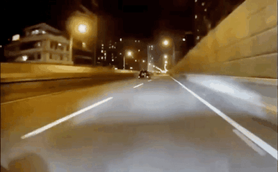 这是一段第一视角拍摄的夜间城市道路驾驶动态图，画面呈现夜晚的城市快速路，道路两侧有建筑物与明亮的路灯，视角跟随车辆向前行驶，前方还有一辆同行车辆，画面随车辆行驶产生晃动，传递出紧张刺激的速度感，仿佛正在进行夜间飙车。