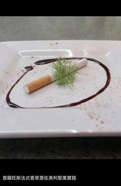 这是一张创意料理摆拍照，白色方形餐盘内，一道模仿万宝路香烟造型的食物被放置其中，搭配普罗旺斯香草叶，用深棕色酱汁在盘内勾勒出圈状，还撒上了调味粉，完全复刻高级法餐的摆盘形式。底部配文「普羅旺斯法式香草葉佐美利堅萬寶路」，玩了谐音与创意料理的反差梗，将香烟转化为“可食用菜品”，用精致法餐的命名方式制造荒诞感，笑点十足。