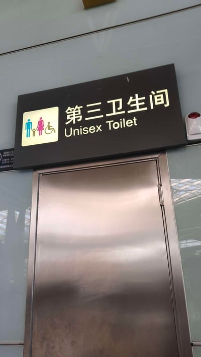 这是一张拍摄于公共场所的第三卫生间标识牌实拍图，标识牌为黑底发光字设计，左侧的发光区域带有包含成人、儿童、轮椅使用者的通用图标，清晰标注了中文“第三卫生间”与英文“Unisex Toilet”。标识牌下方是金属材质的卫生间门，右侧可见一个红色警示装置。第三卫生间是为家庭（携带儿童）、残障人士等特殊人群提供的人性化公共如厕设施，兼顾了不同群体的特殊需求。