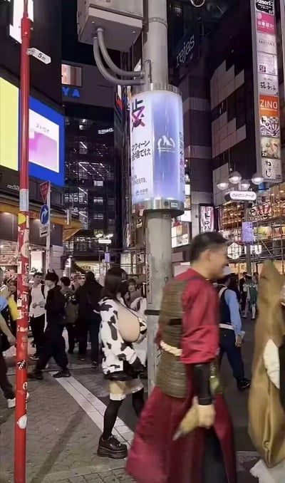 这是夜晚的东京涩谷街头，正值万圣节活动期间，繁华的商圈街道上挤满了人群，不少民众穿着创意十足的cosplay服装参与活动。图中可见一位身着奶牛风格穿搭搭配夸张假胸造型的女生，还有一位穿着红色武士风格服饰的男士正在行走，周围的建筑与招牌凸显出涩谷商圈的标志性特征，整体氛围热闹喧嚣，充满万圣节特有的搞怪与欢快气息。