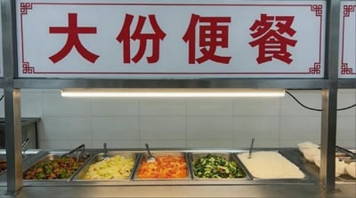 图片展示了一个食堂的取餐区场景。正上方有一块显眼的白色招牌，上面用红色大字写着'大份便餐'，招牌四角有传统的中式花纹装饰。招牌下方是不锈钢的取餐台，台面上并排放置着多个金属餐盆，里面盛放着不同的菜品，包括绿色蔬菜、橙色的番茄炒蛋、白色的米饭等，餐盆上方有一盏长条形的照明灯提供照明。背景是浅色的瓷砖墙面，整体环境整洁，呈现出典型的食堂或快餐取餐区的日常景象。
