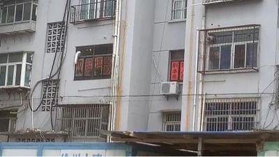这是一张原始拍摄的横版照片，呈现了一栋老式多层居民楼的外观。楼体为浅灰色，外墙上分布着多个带有防盗栏的窗户、空调外机，还有外露的管线，显得颇具年代感。其中一个二层窗户位置悬挂着一块深色招牌，上面写有白色的“汉阴焊部”字样，旁边的窗户上贴着红色的对联，楼体下方还有简易搭建的棚子。整体场景充满生活化气息，是典型的老城区居民楼样貌，图中没有双关、谐音梗或暗示类内容。