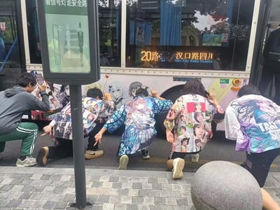 在上海街头，一辆印有《BanG Dream!》《约会大作战》等二次元角色的20路痛车停靠在路边，几位身着印满二次元角色“痛衣”的粉丝，摆出朝拜的姿势对着痛车，另有一位粉丝蹲在一旁用手机记录下这个场景。整体画面充满浓厚的二次元文化氛围，是粉丝对喜爱的二次元作品的狂热应援行为。