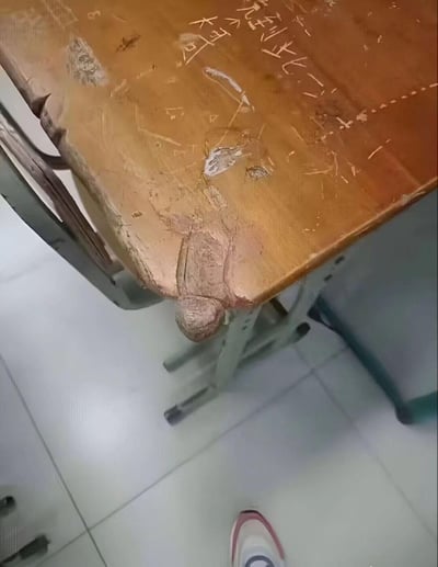 这是一张拍摄于教室的照片，展示了一张使用痕迹严重的旧课桌：桌面漆面多处剥落、磨损，还刻有“走到此一游”“大哥”等手写痕迹，金属桌架也有老化的迹象。最有趣的是桌角的磨损部分因为视角巧合，形成了视觉错位，看起来像一个蜷缩蹲着的人，带来意外的趣味感。

视觉错位说明：桌角的木材磨损痕迹在特定拍摄视角下，轮廓酷似一个蜷缩的人物，属于日常场景里的巧合视觉错觉，并非真实存在的人物。