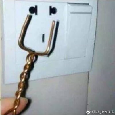 这是一张利用视觉错位效果拍摄的趣味照片：将金属挂链的U型挂钩插进墙壁三孔插座的两个上孔，挂钩的形状和插座的孔洞组合在一起，恰好形成了一个类似吐舌头的搞怪人脸造型，荒诞又充满脑洞，制造出搞笑的视觉错觉效果。