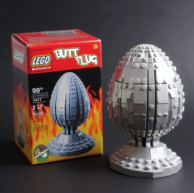 图片展示了一款恶搞乐高（LEGO）产品，左侧为红色包装盒，印有白色“BUTT PLUG”字样及银色蛋形积木模型图案，背景有火焰设计。包装盒上标注产品编号1477、价格99美元、适合年龄3-17岁，以及“OverFucked.com”网址。右侧是已拼装完成的银色蛋形乐高模型，表面由灰色积木块拼接而成，底部有圆形底座。该作品通过模仿乐高官方包装风格，使用不雅产品名称制造幽默讽刺效果，属于粉丝创作的恶搞内容。