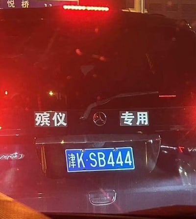 这张图片展示了一辆黑色奔驰品牌的殡仪专用车尾部，车尾中央是奔驰的三叉星徽标志，标志左右两侧分别标注着白色的“殡仪”与“专用”字样，下方悬挂着蓝色的机动车牌照，牌照号码为津K·SB444。
车牌号存在明显的谐音与数字暗示梗：“SB”在中文网络语境中常作为脏话的谐音使用，数字“444”的发音和中文里的“死”相近，而这辆车恰好是殡仪专用车，车牌号的内容与车辆用途形成了戏剧性的巧合搭配，带有戏谑的趣味感。