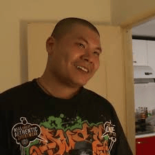 图片中是一位光头男子，穿着印有彩色涂鸦风格文字和图案的黑色T恤，面带微笑看向一侧。背景可见厨房场景，有白色橱柜和红色台面，整体氛围轻松日常。