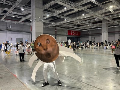 这是在上海国家会展中心的漫展场馆内（背景可见“5.1馆”标识），一位爱好者自制道具cos了《间谍过家家》里阿尼亚脑补的“花生人”造型：棕色圆形的花生头套开了两个洞露出脸，搭配白色紧身裤和自制的白色“花生须”肢体，在漫展现场摆出姿势。场馆内还有不少其他漫展参与者、专业摄影设备，整体氛围热闹。这个cos精准还原了动画里阿尼亚的经典脑洞场景，造型搞笑吸睛，成为现场的焦点之一。