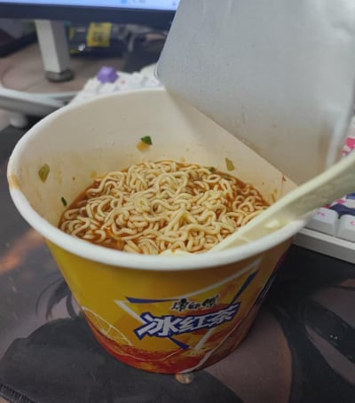 这是一张办公桌面的实拍图，画面核心是一个康师傅冰红茶的纸杯，里面盛放着泡好的带红油汤汁的方便面，还插着一把塑料泡面叉，杯子的盖子被掀开至一侧。背景中可以看到部分机械键盘和电脑显示器，呈现出典型的工位用餐场景。这是一种非常随性的凑活用式餐食操作，没有双关、谐音梗或特殊暗示。