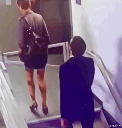 这是一段趣味动态GIF，画面中一位身着黑色短裙、高跟鞋的女士正走上自动扶梯，在她身后的视角下，扶梯台阶与女士腿部形成了巧妙的视觉错位，看起来女士的腿部“消失”了，只剩上半身和脚上的鞋子，制造出类似灵异的滑稽错觉，实际是拍摄角度带来的巧合，让人忍俊不禁。