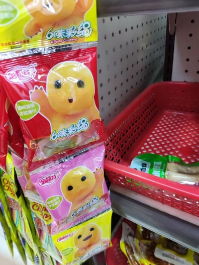 这张图是超市货架区域的实拍照片，画面左侧竖直堆叠着多袋金稻谷品牌的6味彩绳软糖，包装袋有红色、粉色、黄色三种不同的底色，每袋包装上都印有黄色的奶龙卡通形象，还标注了“边吃边玩才过瘾!!!”的宣传语，软糖旁还能看到珍宝珠等其他品牌的零食；画面右侧是放置在货架上的红色镂空塑料购物篮，篮内装有其他零食，背景为超市常用的洞洞板货架结构。