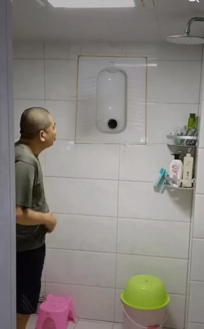 这张图是拍摄于家庭卫生间内的趣味错觉照片，卫生间墙面铺着白色瓷砖，一名留寸头的中年男子站在画面左侧，身体侧向墙面，正一脸诧异疑惑地盯着墙上方一块被透明玻璃框起来的区域；这块区域内的白色长形造型搭配下方的黑色圆点，外形和嵌入式安装的小便池几乎一模一样，实际上这只是墙面改造后留下的装饰造型，并不是真的小便池。画面左下角摆放着一个粉色塑料小凳子，右下角是一个带亮绿色盖子的水桶，画面右侧墙面安装了置物架，上面摆放着各类洗浴用品，右上角可见淋浴顶喷，整体是因视觉错觉引发的搞笑场景。