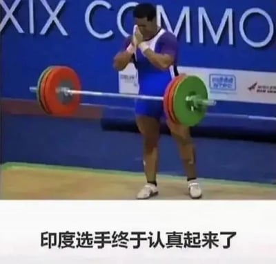 图片中是一位身着蓝紫色运动服的印度举重选手，正站在举重比赛的赛场中，双手合十放在脸前，似乎在进行赛前的专注准备。他身后是蓝色的背景板，上面能看到部分白色字母“COMMO”。图片底部配有文字“印度选手终于认真起来了”，整体场景展现了运动员在比赛中专注投入的瞬间。