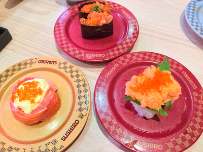 这是一张在寿司郎（SUSHIRO）门店内拍摄的原始实拍图，浅木色的餐桌上摆放着三个印有“SUSHIRO”品牌标识的回转寿司专用盘：其中金色格纹盘上盛放的是三文鱼卷寿司，表面铺有浅黄色沙拉酱和橙色飞鱼籽；上方的红色格纹盘里是海苔包裹的三文鱼碎军舰寿司，顶部点缀有绿色葱花；右下角的红色格纹盘里是三文鱼泥搭配飞鱼籽的寿司，底部为白米饭，还搭配了紫苏叶作为装饰，整体呈现出日式回转寿司的用餐场景，餐品色泽鲜亮诱人。