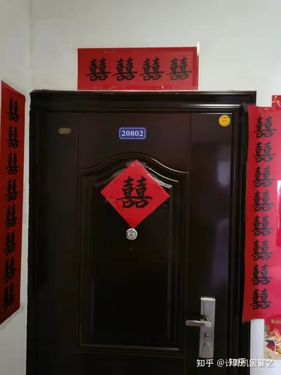 这是一张拍摄居民楼入户门的实拍照片，门牌号为20802的深棕色入户门及周边被布置了多处红底黑字的囍字：门正中央贴有菱形的单囍，门上方横贴四个并排的囍字，门的左右两侧各竖贴一列多个囍字，整体是中式婚礼的婚房布置风格，充满浓厚的喜庆氛围，图中没有双关、谐音梗或暗示内容。