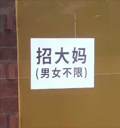 图片中展示了一张贴在棕色墙壁上的白色招聘启事，左侧可见部分红砖墙。启事上用黑色字体写着“招大妈”，下方括号内标注“（男女不限）”。内容存在明显矛盾：“大妈”通常指中老年女性，却又注明“男女不限”，形成一种幽默的反差感，让人忍俊不禁。