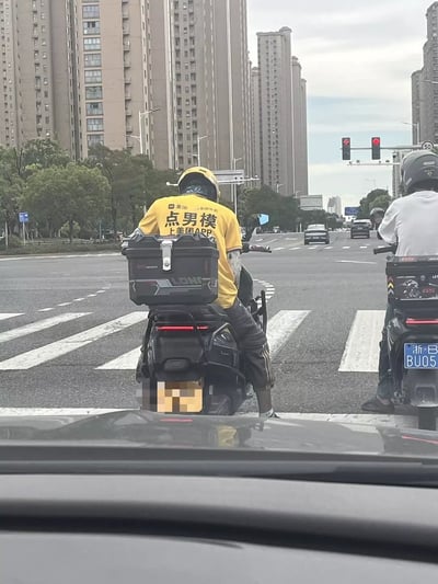 整张图是从机动车驾驶室内向前拍摄的白天城市道路路口场景，路口交通信号灯显示为红灯。画面正中是一名外卖配送员，佩戴黄色头盔、身穿黄色美团风格工服，正骑电动车停在斑马线前等待通行，他的工服后背印着被恶搞的标语“点男模 上美团APP”，替换了原本的美团外卖官方宣传语，反差感强烈十分搞笑。外卖员左侧还有另一名佩戴灰色头盔、穿白色上衣的电动车骑手也在等待通行，背景可见道路两侧的绿树、连片的高层住宅楼宇，远处还有其他行驶的机动车。画面最下方露出了拍摄车辆的前挡风玻璃下沿和中控台顶部。