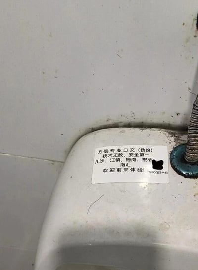 这是一张在公共卫生间小便池区域拍摄的照片，画面中的小便池边缘贴着一张低俗不良的小广告贴纸。贴纸内容标注“无偿专业口交（伪娘）”，附带宣传语“技术无敌、安全第一”，列出了川沙、江镇、施湾、祝桥、南汇这些上海下属区域，还有被打码处理的QQ二维码，文字引导观看者“打开QQ扫一扫”“欢迎前来体验”。拍摄场景的卫浴设施和墙面有明显污渍，整体环境显得脏乱，而贴纸内容属于违反公序良俗的低俗色情性质违规宣传内容。