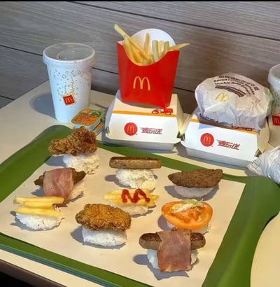图中展示了一份麦当劳麦乐送套餐，包含薯条、饮料、培根牛肉双层堡、麦乐鸡等餐品，在前方的托盘上，创作者将麦当劳的食材（炸鸡块、培根、牛肉饼、香肠、薯条等）搭配米饭，摆放成日式寿司的样式，玩起了麦当劳与寿司郎的联动梗，把西式快餐改造成日式料理的形态，充满创意与趣味性，是一种脑洞大开的美食DIY展示。