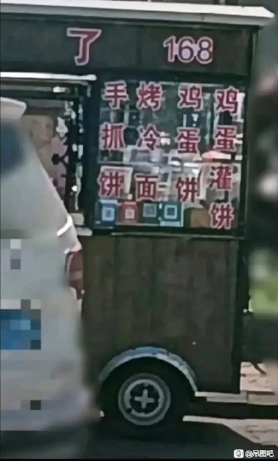 这是一张竖屏实拍的街头移动小吃车图片，餐车为深色外观，底部带有轮子。餐车窗口上方的红色文字分为两部分，左侧是单独的“了”字，右侧是数字“168”；窗口区域的菜单采用四列三行的竖排文字布局，竖读依次为手抓饼、烤冷面、鸡蛋饼、鸡蛋灌饼，这种横排的文字排列方式会让初次观看的人产生阅读混淆，形成趣味效果。餐车窗口内可见一位模糊的真人摊主，画面左侧有白色车辆部分遮挡餐车，图片右下角带有“@吊图吧”的水印标识。