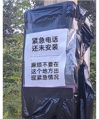 这是一张户外原始拍摄的静态照片，画面主体是一个被黑色塑料布包裹的未启用紧急电话装置，装置表面贴着一张白色提示纸条。纸条分为上下两部分，上方标注“紧急电话还未安装”，下方用幽默无奈的语气写着“麻烦不要在这个地方出现紧急情况”，以一种反常规的提示方式，表达了此处紧急电话无法使用的现状，充满了搞笑的反差感，让严肃的提示变得趣味十足。