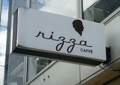 这是一张实地拍摄的Rizza CAFFÈ咖啡店户外招牌照片，招牌为悬挂式白色灯箱，带有深色边框，安装在现代玻璃幕墙建筑外墙上。招牌上采用艺术化的手写风格字体呈现品牌名“rizza”，右侧配有意大利语的“CAFFÈ”字样，同时在品牌名上方有一个深色的人物侧脸剪影，整体视觉设计简洁精致，没有双关、谐音梗或暗示内容。