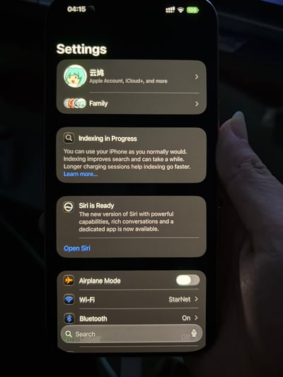 这是一张真人手持iPhone拍摄的手机设置界面实拍图，界面顶部显示时间为04:15，电量100%。最上方是Apple账户区域，用户名为“云鸠”，搭配绿发卡通形象头像，下方是家庭功能入口；接下来有两条系统提示：第一条是索引进行中的提示，告知用户可正常使用手机，索引可优化搜索速度，长时间充电能加快索引进度，附带了解更多入口；第二条是新版Siri就绪提示，说明新版Siri具备更强功能、更丰富的对话能力且有专属应用，附带打开Siri的入口；下方依次是飞行模式（当前处于关闭状态）、Wi-Fi（已连接名为StarNet的网络）、蓝牙（已开启）的设置选项，最底部是系统搜索栏。