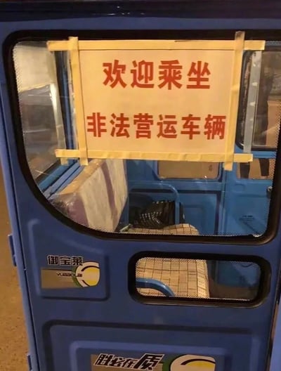 这是一张原始拍摄的静态照片，主体是一辆蓝色的“御宝莱”牌电动载客三轮车。在车辆的侧窗上，用黄色胶带固定着一张白底红字的标语，内容为“欢迎乘坐非法营运车辆”，直白地自曝了该车辆属于非法营运的性质，形成了强烈的反差感与趣味性。车门左下方印有“御宝莱 YUBAOLAI”的品牌标识，车门底部还有“胜必在质”的字样，透过车窗可以看到车内简单的座椅设施，整体是日常街头的真实场景。