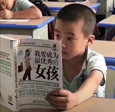 这是一张拍摄于小学教室的静态实拍图，画面主体是一位短发小男孩，他坐在蓝色边框的课桌前，正双手捧着一本名为《我要成为最优秀的女孩》的励志书籍专注阅读，男孩的神情认真投入。背景中可以看到其他同学的课桌和趴在桌上的同学，还原了真实的课堂场景。男孩作为男性阅读面向女孩的励志书籍，这种场景上的错位形成了强烈的反差感，让画面充满了滑稽搞笑的氛围。书籍封面上还标注了副标题“让女孩拥有完美性格的70个故事”，进一步凸显了这种内容与读者的反差。