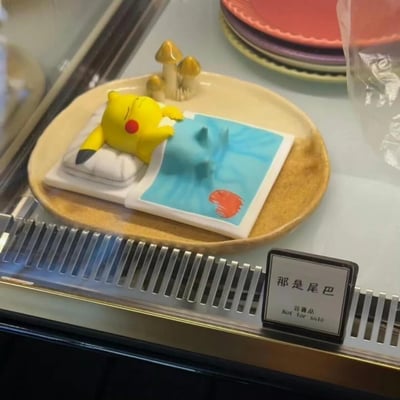 这是一款宝可梦主题的创意展示摆件，画面中皮卡丘正躺在床上安睡，盖着的被子被设计成立体的杰尼龟背部造型，制造出“皮卡丘把杰尼龟当被子盖”的趣味视觉错觉。旁边的标识牌标注着“那是尾巴 非卖品 Not for sale”，直接点明这个搞笑设计的真相，让这个摆件充满趣味，十分戳中宝可梦粉丝的笑点。