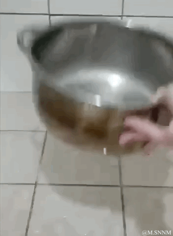 这是一段动态GIF，画面中一只手握着不锈钢锅的底部，由于角度和姿势的巧合，锅身的棕色区域和手的肤色、位置形成了视觉错位，看起来仿佛锅“咬”住了手，或者手和锅的下半部分融合在了一起，制造出荒诞又搞笑的视觉错觉效果。