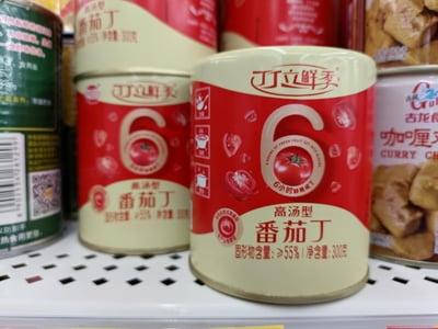 这是一张超市货架的实拍图，主体是**丁立鲜季高汤型番茄丁罐头**，罐头采用红米配色包装，标注了「6小时鲜果成丁」「固形物含量≥55%」「净含量300克」的产品信息，还附带了炒菜、焖煮、煲汤的烹饪场景提示。罐头周围还摆放着其他品牌的食品罐头，右侧是古龙的咖喱罐头，左侧是绿色包装的罐头，呈现出日常超市的食品售卖场景。