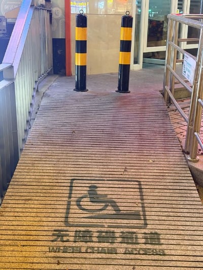 这是一张实拍的公共场景照片，画面主体是一条铺设有防滑条纹的无障碍坡道，坡道表面印有标准的轮椅图案标识、“无障碍通道”汉字及对应的英文标注，坡道两侧分别设置了金属扶手，通往后方的商铺入口。但在坡道的顶端、入口位置竖直设立了两根黑黄相间的防撞隔离柱，两根柱子的间距不足以让轮椅通行，直接阻断了无障碍通道的通行路径，让无障碍设施完全失去了应有的作用，属于不合理的公共设施设置案例。