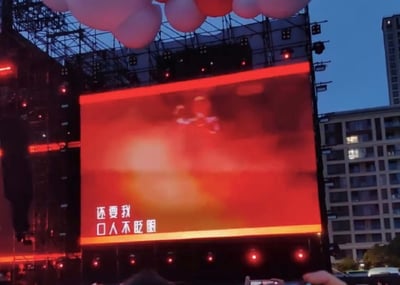 这是一张户外音乐节现场的拍摄图，画面主体是舞台上的大型电子屏幕，屏幕底色为红色，上面有模糊的演出人物形象，同时显示着“还要我 口人不眨眼”的白色文字，这里是将“杀”字拆分为“口人”，玩了拆字的谐音梗，替代了原句“还要我杀人不眨眼”，营造出戏谑的效果。背景能看到舞台的金属灯光架和旁边的居民楼，现场氛围是演出进行中的热闹场景。