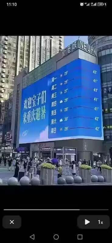 图片展示了重庆繁华商圈的巨型电子广告牌，屏幕上以蓝色为底色，显示着一周天气预报和欢迎标语。画面中可见高楼林立的城市背景，广告牌下方有许多行人正在街道上行走，周围有绿植和装饰性球体。广告牌左侧写着“欢迎主子们来重庆避暑”，右侧则是周一至周日的温度信息，最高温度在39°到27°之间波动。整体呈现出重庆作为热门旅游城市的热闹氛围和夏季气候特点。