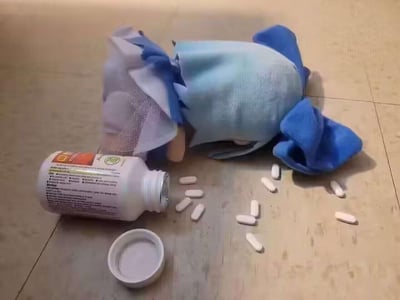 这是一张充满脑洞的视觉错位创意图，利用蓝色口罩和布料拼接摆放，制造出一个“倒地的人”的视觉假象；旁边的药瓶被打翻，白色药片散落在地面，进一步营造出“服药后晕倒”的逼真场景。实际上是用日常物品的巧妙摆放制造的趣味错觉，诙谐又有创意。